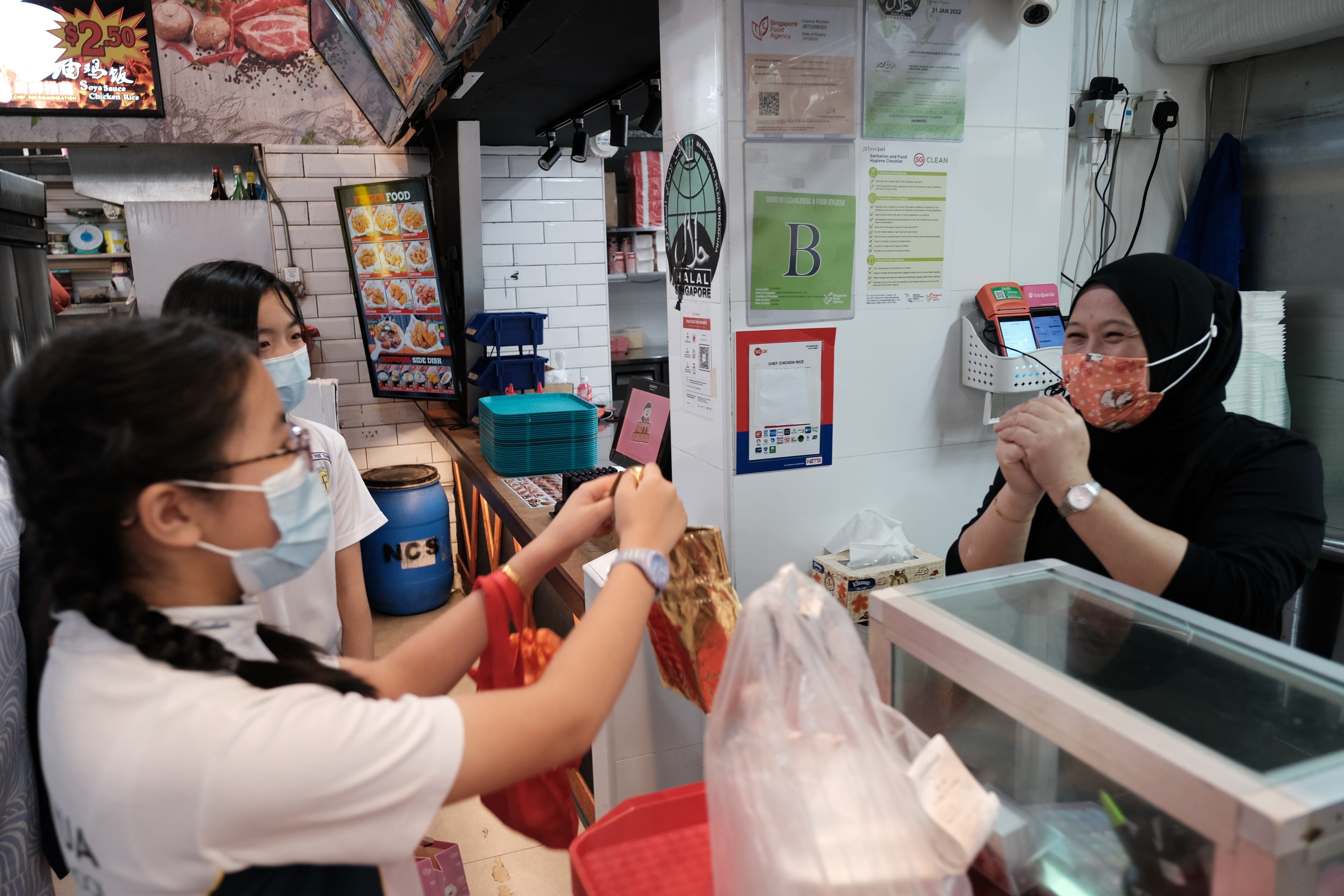 Two masked girls order food from a masked vendor at a Halal-certified Singapore hawker stall priced at $2.50.